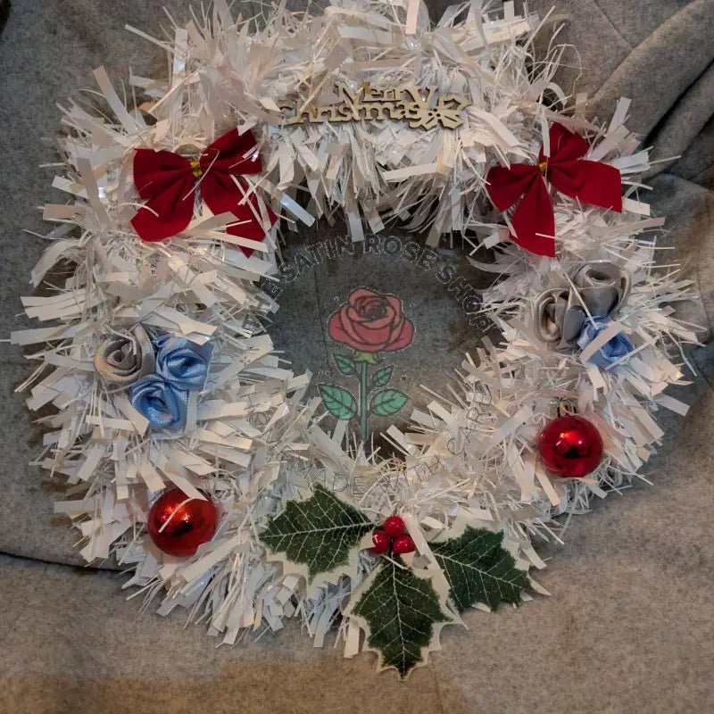 Decorated satin rose Christmas wreath with white tinsel, red bows, silver and blue roses, red baubles, and green holly leaves